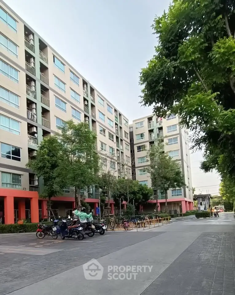 Modern apartment building exterior with lush greenery and parked motorcycles in urban setting.