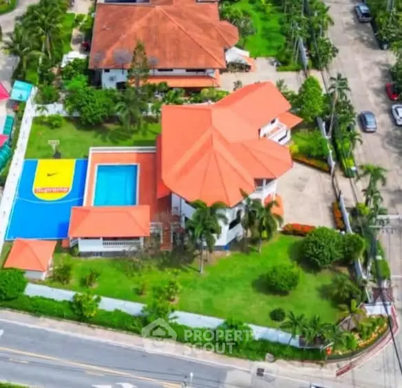 Stunning aerial view of luxury villa with pool and vibrant sports court, surrounded by lush greenery.