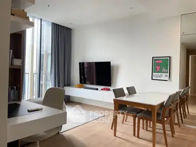 Modern living room with sleek furniture and large TV, featuring a dining area and natural light from tall windows.