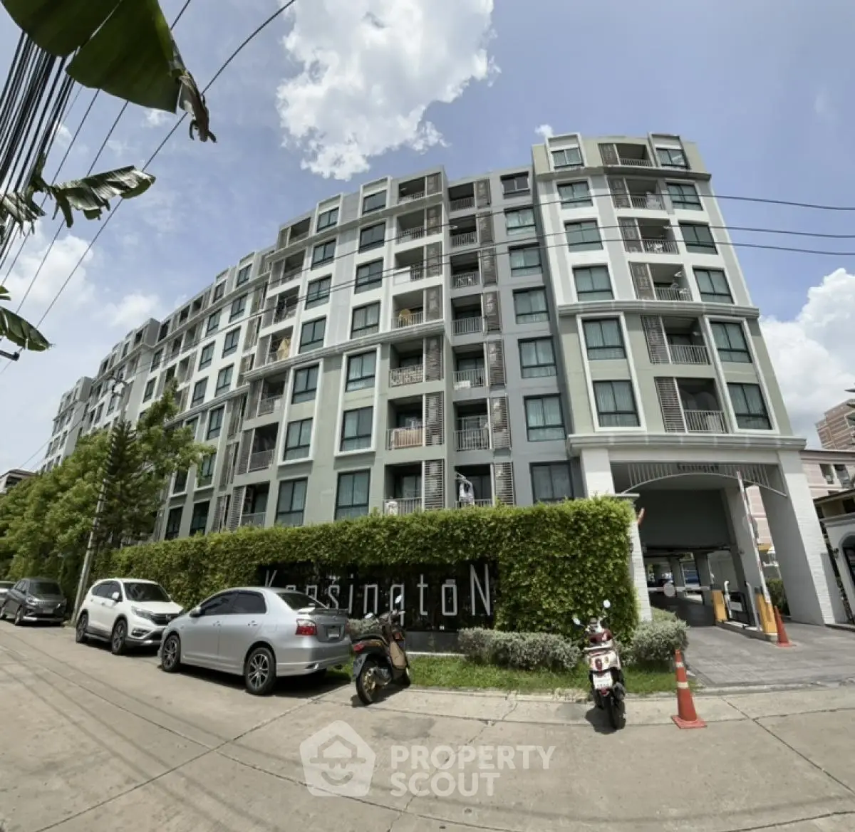 Modern multi-story residential building with lush greenery and parked vehicles in urban setting.