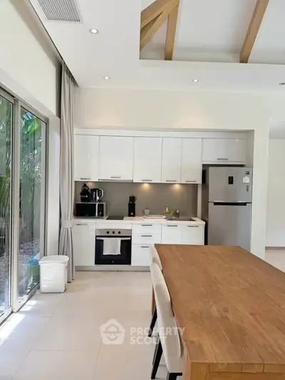 Modern kitchen with sleek white cabinets and wooden dining table, featuring large windows and high ceilings.
