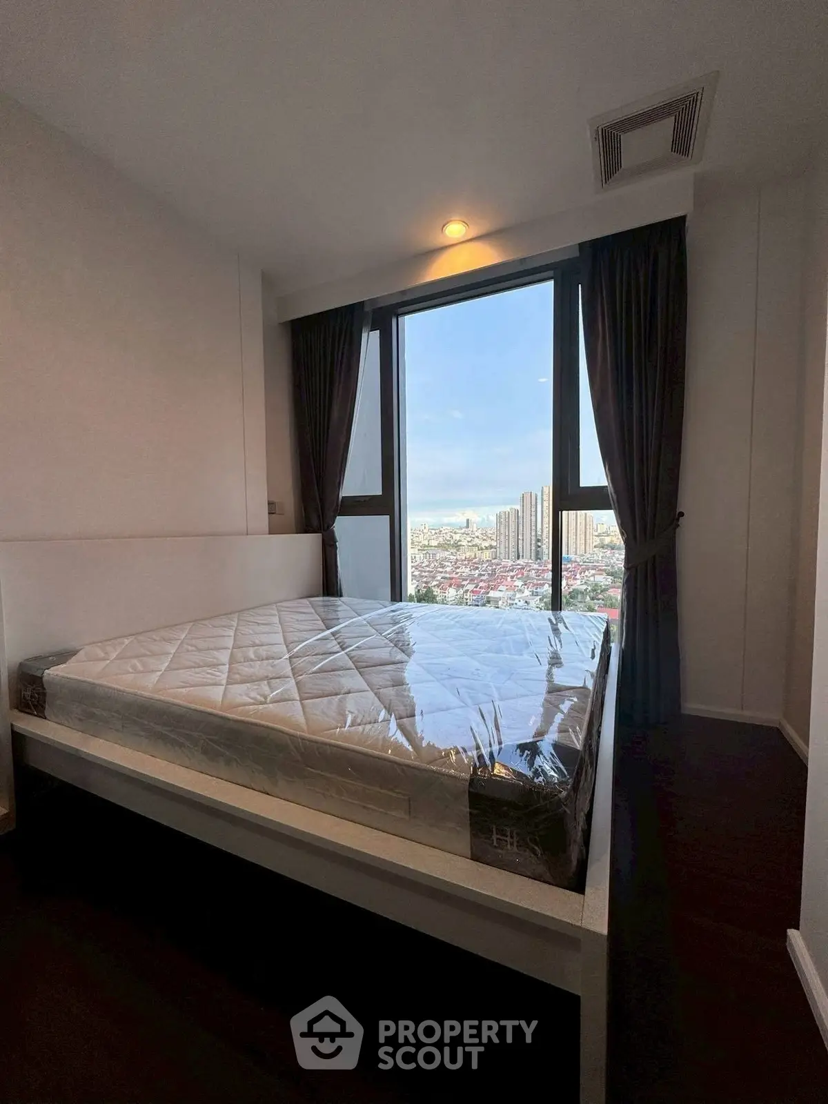 Modern bedroom with large window offering stunning city view, featuring a new mattress and elegant dark curtains.