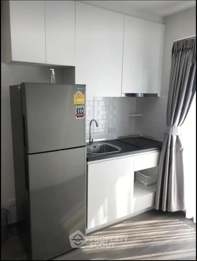 Modern compact kitchen with sleek white cabinets and stainless steel fridge in bright apartment.