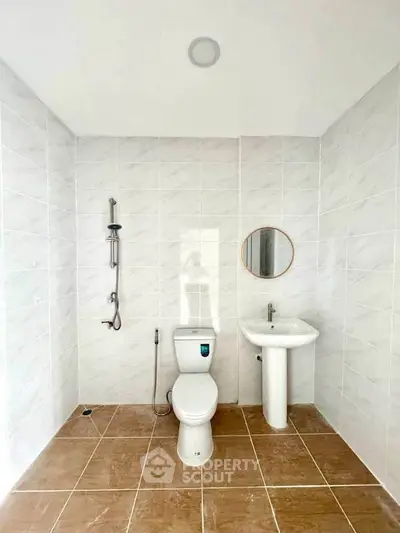 Modern minimalist bathroom with white tiles and brown floor, featuring a toilet, sink, and shower.