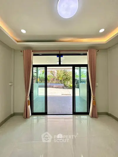 Spacious living room with large glass doors opening to a garden view, featuring elegant curtains and modern lighting.