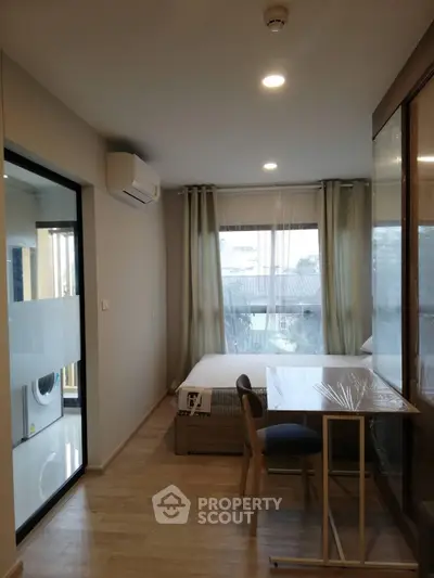 Modern studio apartment with wooden flooring and large window, featuring a compact dining area and visible washing machine.