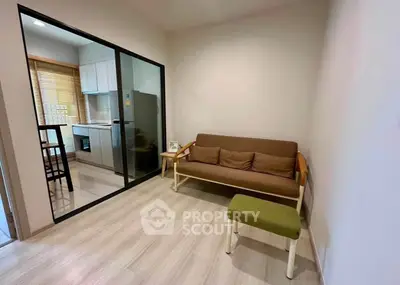 Modern living room with glass partition leading to a compact kitchen, featuring sleek furniture and wooden flooring.