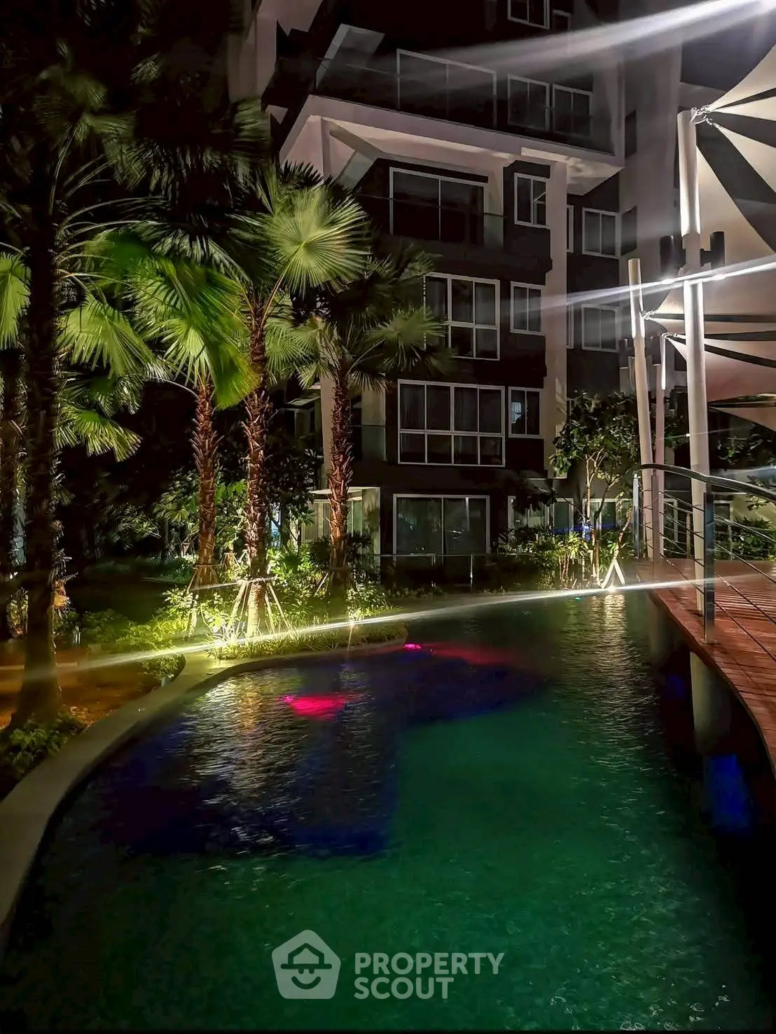Luxurious night view of a modern apartment building with illuminated pool and lush greenery.