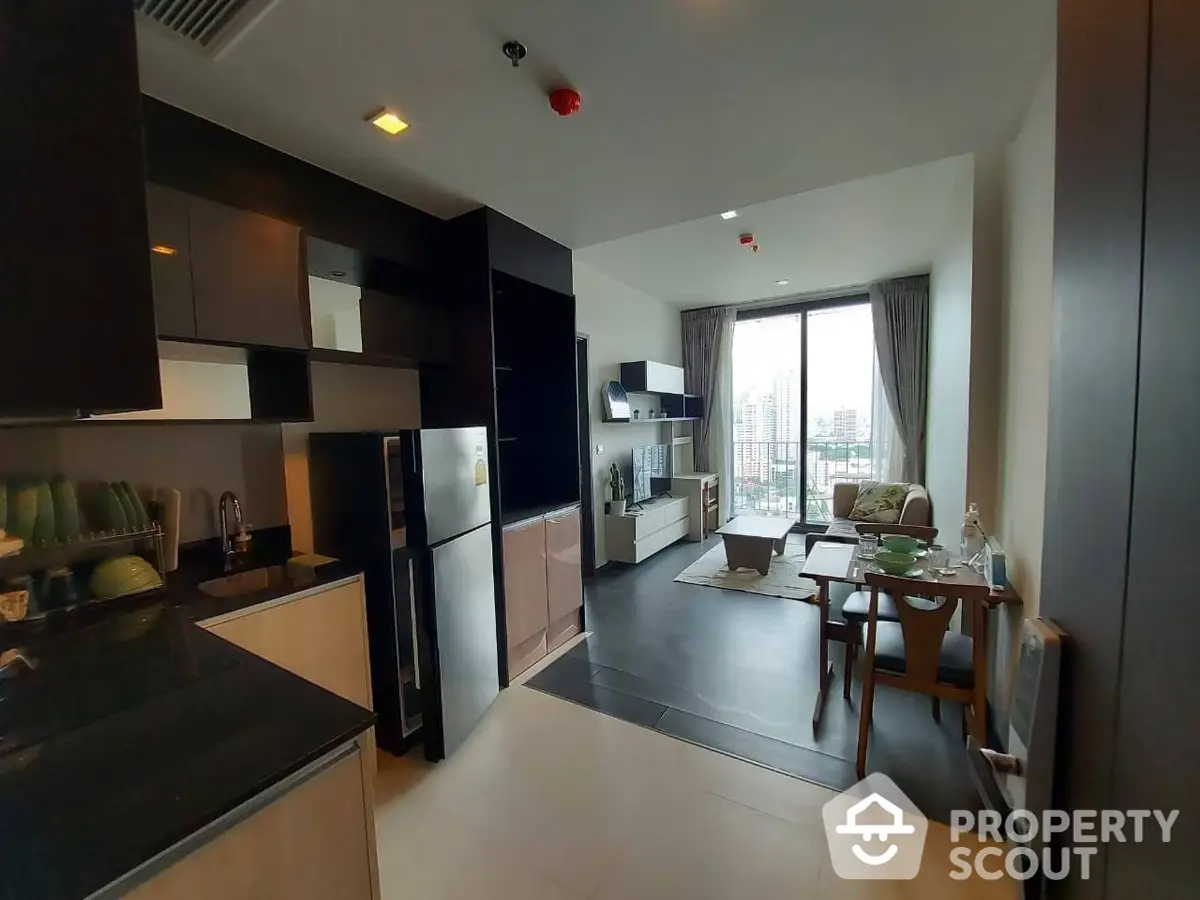 Modern open-plan kitchen with sleek black cabinetry flows into a cozy living area, bathed in natural light from the large window offering a cityscape view.