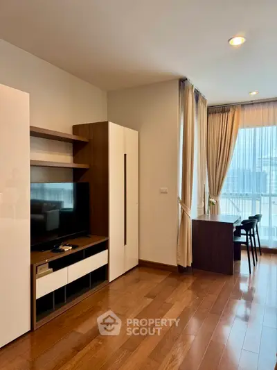 Modern living room with wooden flooring, TV unit, and large windows offering natural light.