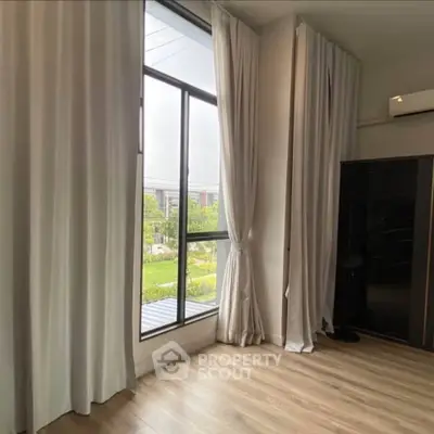 Spacious room with large window and elegant curtains overlooking greenery, featuring sleek wooden flooring.