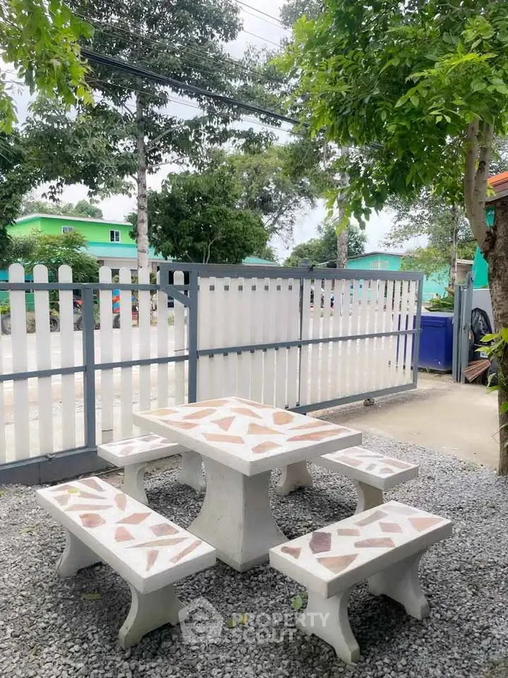 Charming outdoor seating area with stone benches and table, surrounded by lush greenery and a white picket fence.