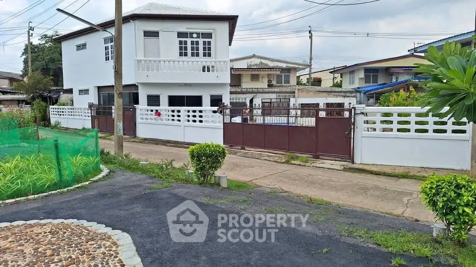 Charming suburban house with white facade and gated entrance, surrounded by lush greenery and a paved driveway.