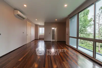 Spacious empty room with polished wooden floors and large windows offering lush green views.