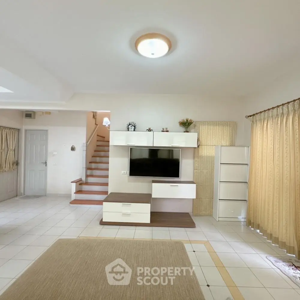 Spacious living room with modern entertainment unit, tiled flooring, and elegant staircase leading to upper level, bathed in natural light from large windows.