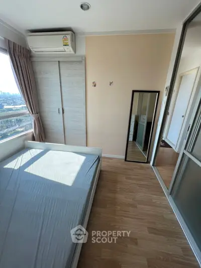Bright bedroom with large window and wooden floor, featuring a wardrobe and mirror.