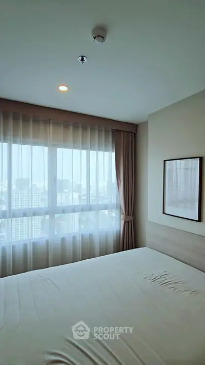 Bright bedroom with large windows and city view, featuring elegant curtains and framed artwork.