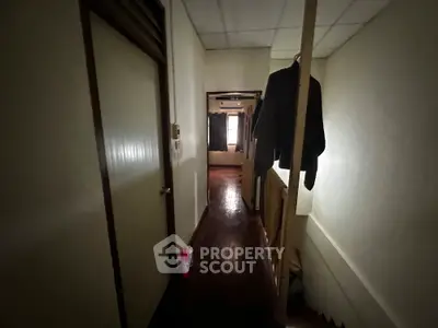 Dimly lit hallway with wooden flooring and hanging clothes, leading to a room with natural light.