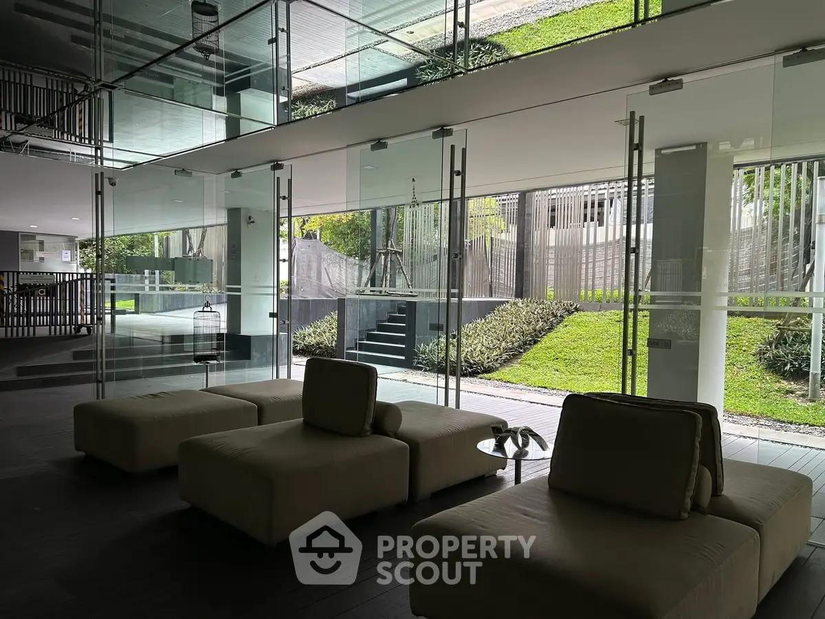 Modern building lobby with glass walls and garden view, featuring stylish seating area.