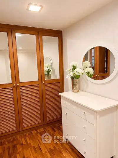 Elegant bedroom with wooden wardrobe and white dresser, featuring a round mirror and floral decor.
