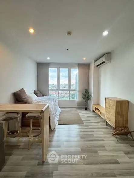 Modern studio apartment with wooden flooring, cozy bed, and city view through large windows.