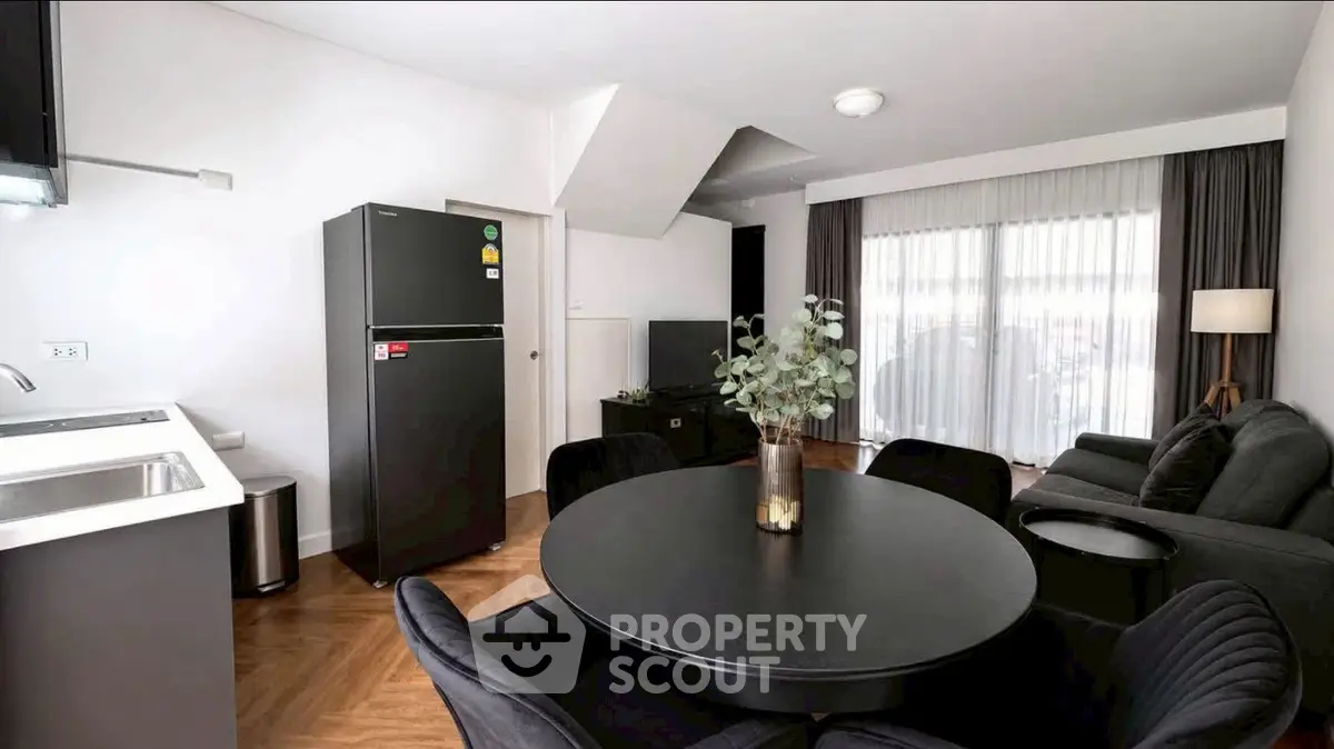 Modern living room with sleek black furniture and open kitchen layout, featuring a stylish dining area and large window.