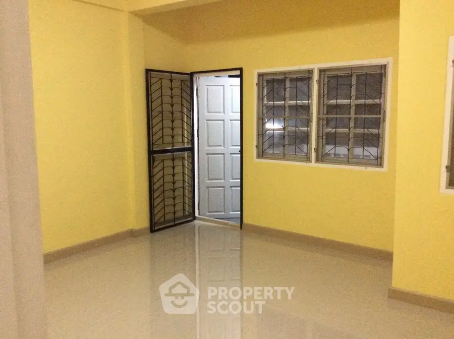 Spacious room with yellow walls and tiled floor, featuring a secure metal door and large windows.