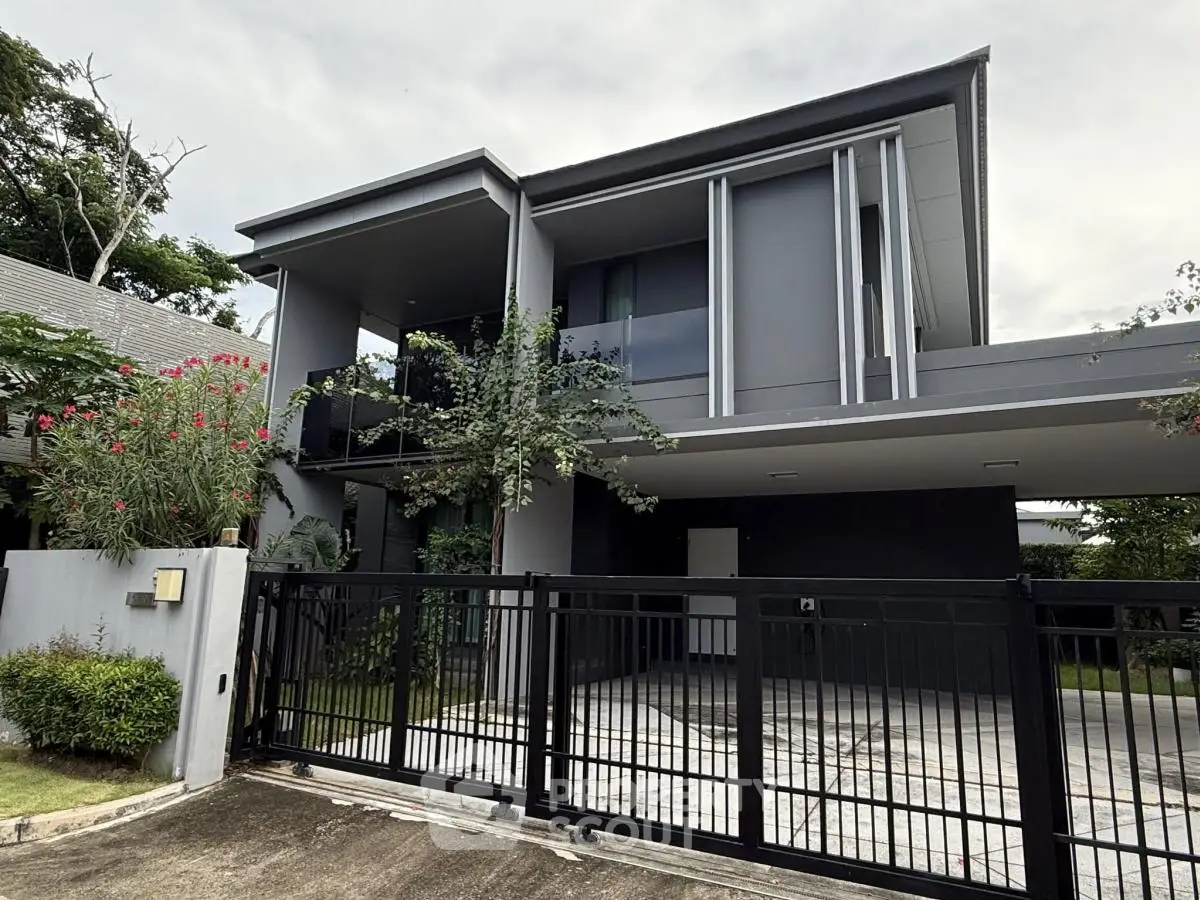 Modern two-story house with sleek design and gated entrance, surrounded by lush greenery.