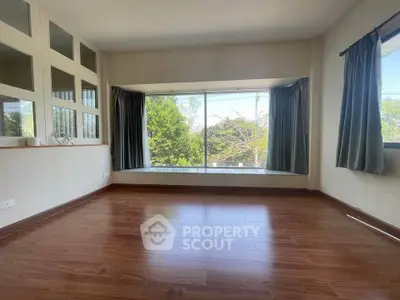 Spacious empty room with large windows and wooden flooring, offering a serene view of lush greenery.