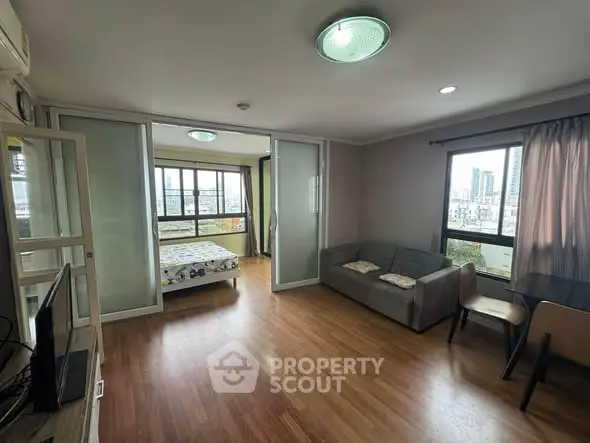 Spacious living room with wooden flooring and city view, featuring a cozy sofa and sliding glass doors to bedroom.