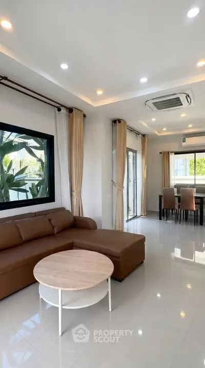 Spacious modern living room with elegant brown sofa and dining area, featuring large windows and stylish decor.