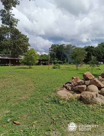 Expansive green lawn with trees and rustic buildings under a cloudy sky, perfect for serene living.