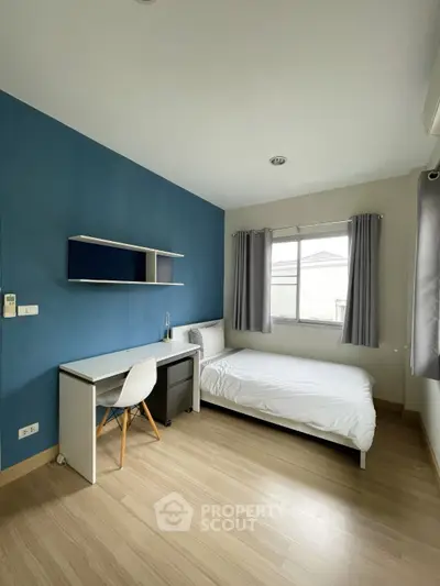 Modern bedroom with blue accent wall, sleek desk, and cozy bed in bright natural light.