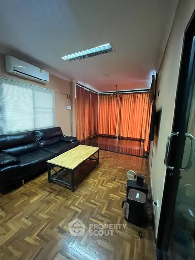 Spacious living room with parquet flooring and modern furnishings, featuring a cozy black leather sofa and large windows.