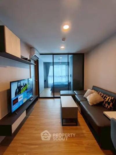 Modern living room with sleek design and large TV, leading to a cozy bedroom through glass doors.