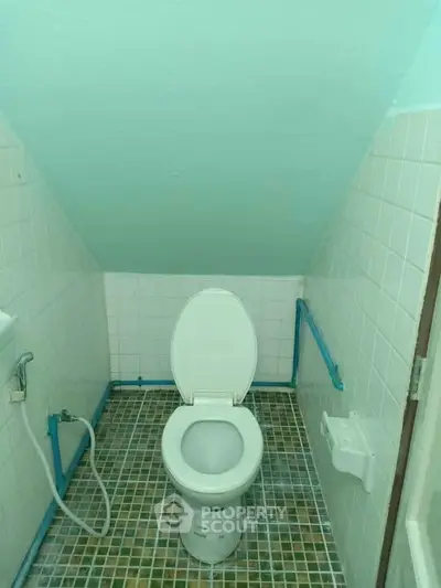 Compact bathroom with sloped ceiling and tiled floor, featuring a toilet and bidet spray.