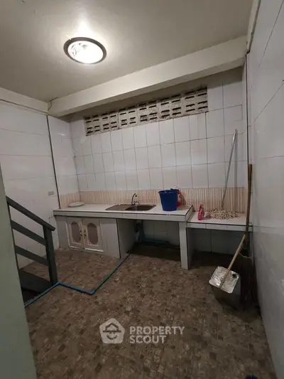 Basic kitchen area with tiled walls and floor, featuring a sink and cleaning supplies.