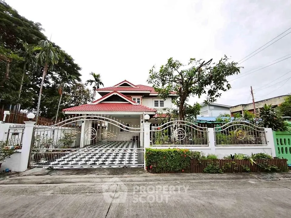 Charming two-story house with red roof and gated driveway in lush suburban setting.