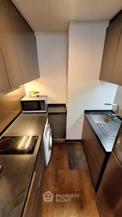 Modern kitchen with sleek dark cabinetry and built-in appliances, featuring a compact layout and wooden flooring.
