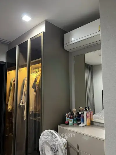 Modern bedroom with sleek wardrobe and air conditioning unit, featuring a dresser with cosmetics and a fan.