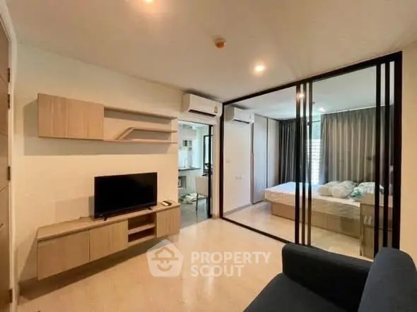Modern living room with glass partition leading to a cozy bedroom, featuring sleek furniture and warm lighting.
