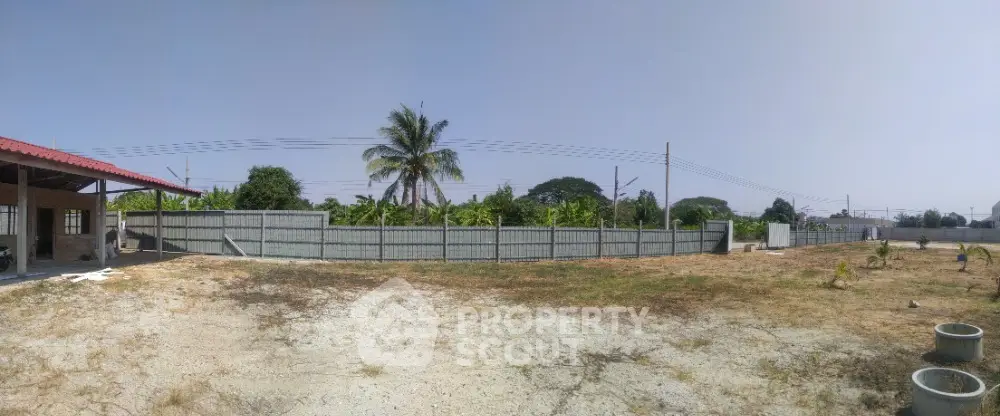 Expansive vacant land with potential for development, surrounded by a secure fence and lush greenery, under a clear blue sky.