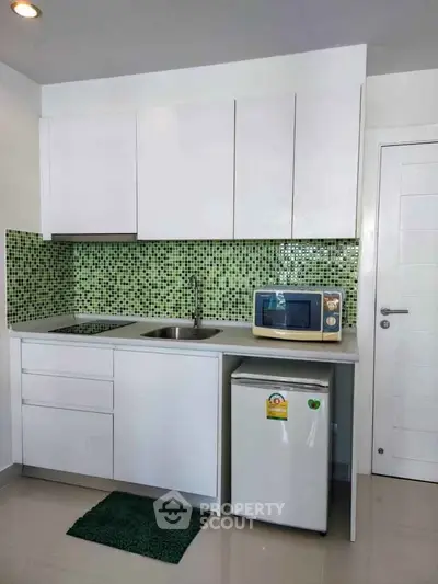 Modern compact kitchen with white cabinets and green mosaic backsplash, featuring a microwave and mini fridge.