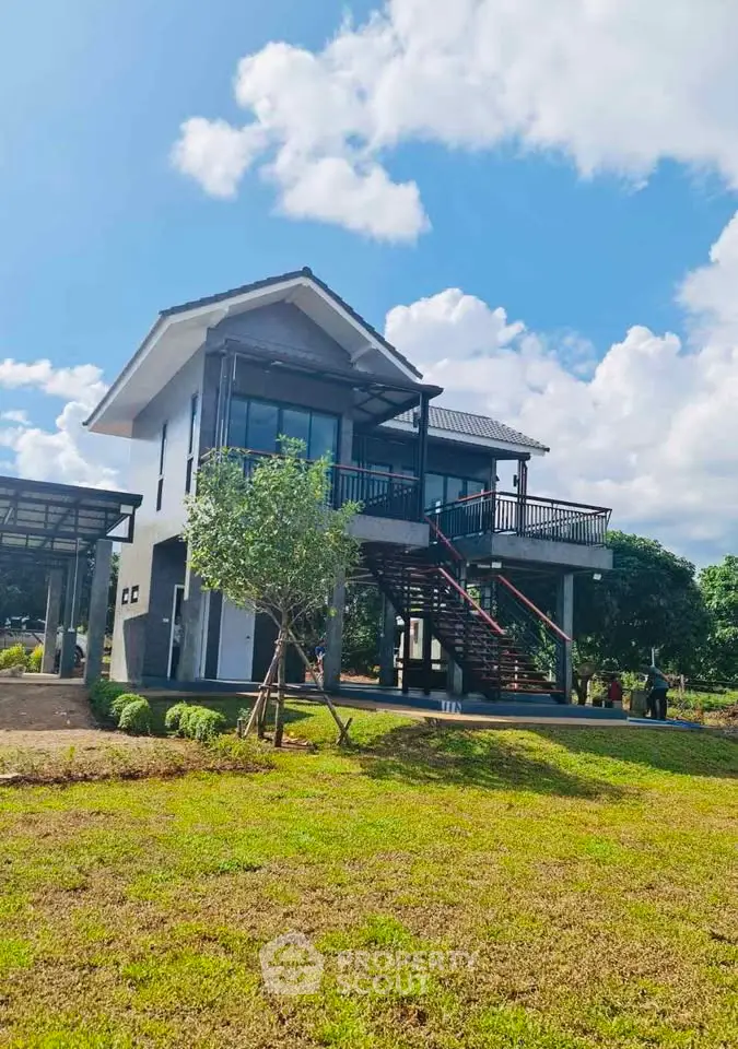 Charming two-story modern house with spacious balcony and lush green surroundings under a clear blue sky.