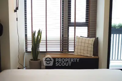 Cozy bedroom with modern decor, featuring a large window with blinds, a potted plant, and a cushioned bench.