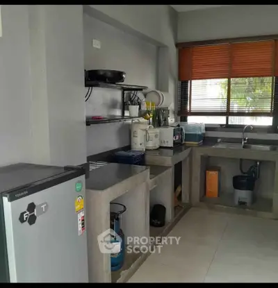 Modern kitchen with concrete countertops and open shelving, featuring a compact fridge and gas stove.