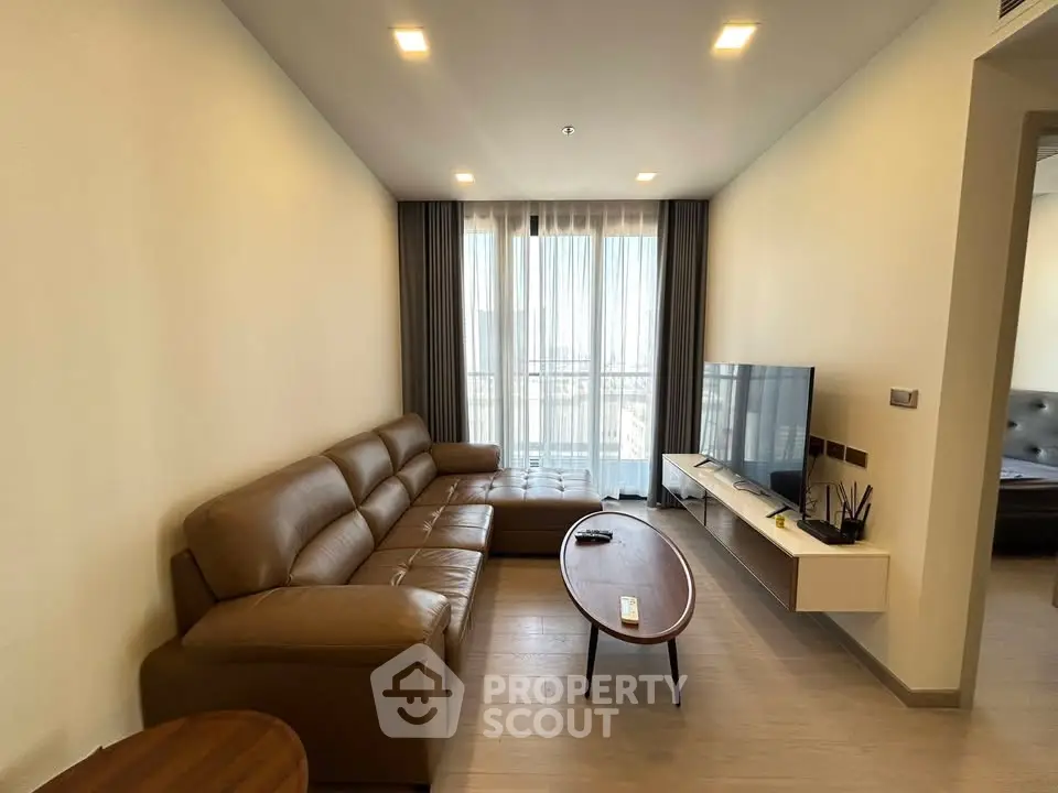 Modern living room with leather sofa and wall-mounted TV, featuring natural light from large window.