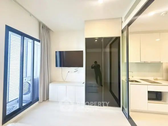 Modern apartment interior with sleek kitchen and wall-mounted TV, featuring large windows for natural light.