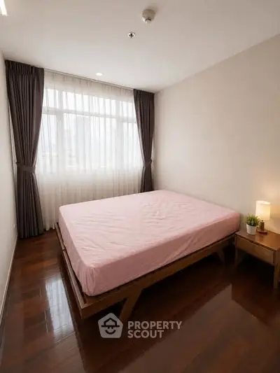 Cozy bedroom with wooden flooring and large window with curtains, featuring a neatly made bed and bedside table.