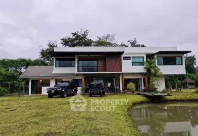 Modern two-story house with large windows and lush garden, featuring a serene pond and parked vehicles.